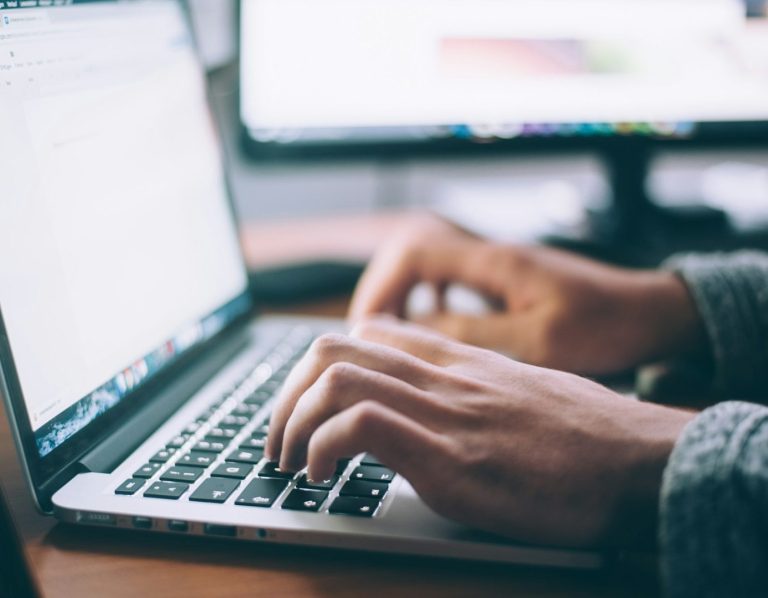 Person typing on a laptop keyboard with a blurred background of another monitor.